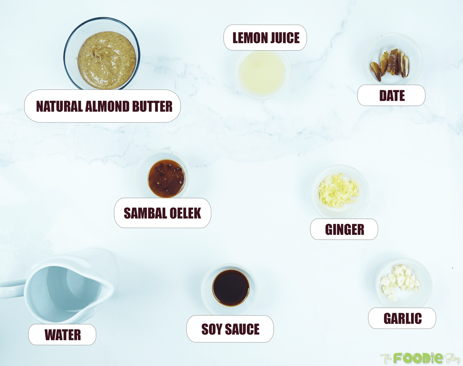 Overhead view of almond butter, lemon juice, tamari, garlic, ginger, sambal oelek, date, and water labeled on a white surface.