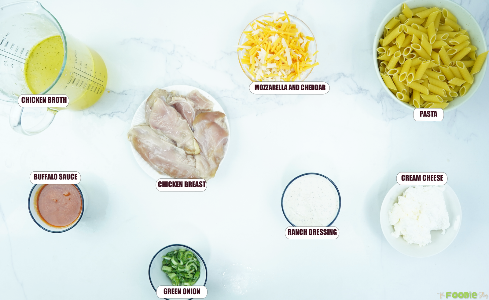 Ingredients for Crock Pot Buffalo Chicken Pasta arranged on a white surface, including chicken breast, penne, Buffalo sauce, ranch, cream cheese, broth, cheese, and green onions.