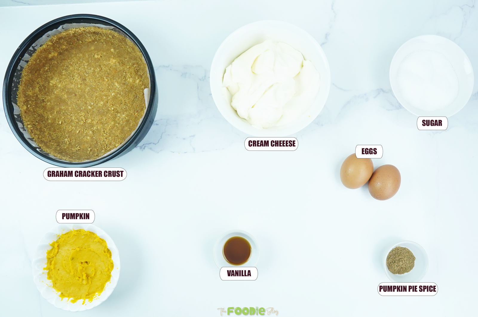 Ingredients for Easy Pumpkin Pie Cheesecake arranged on a white marble surface, including graham cracker crust, cream cheese, eggs, sugar, pumpkin puree, vanilla, and pumpkin pie spice.