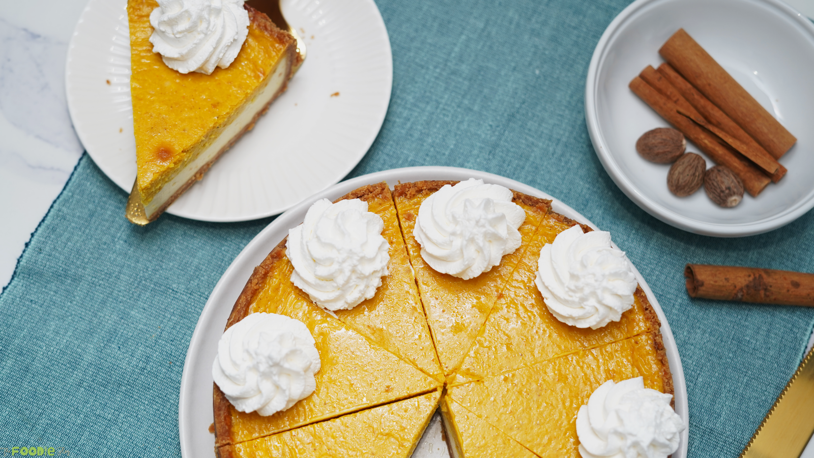 A sliced pumpkin pie cheesecake topped with whipped cream on a white plate with cinnamon sticks and nutmeg nearby.