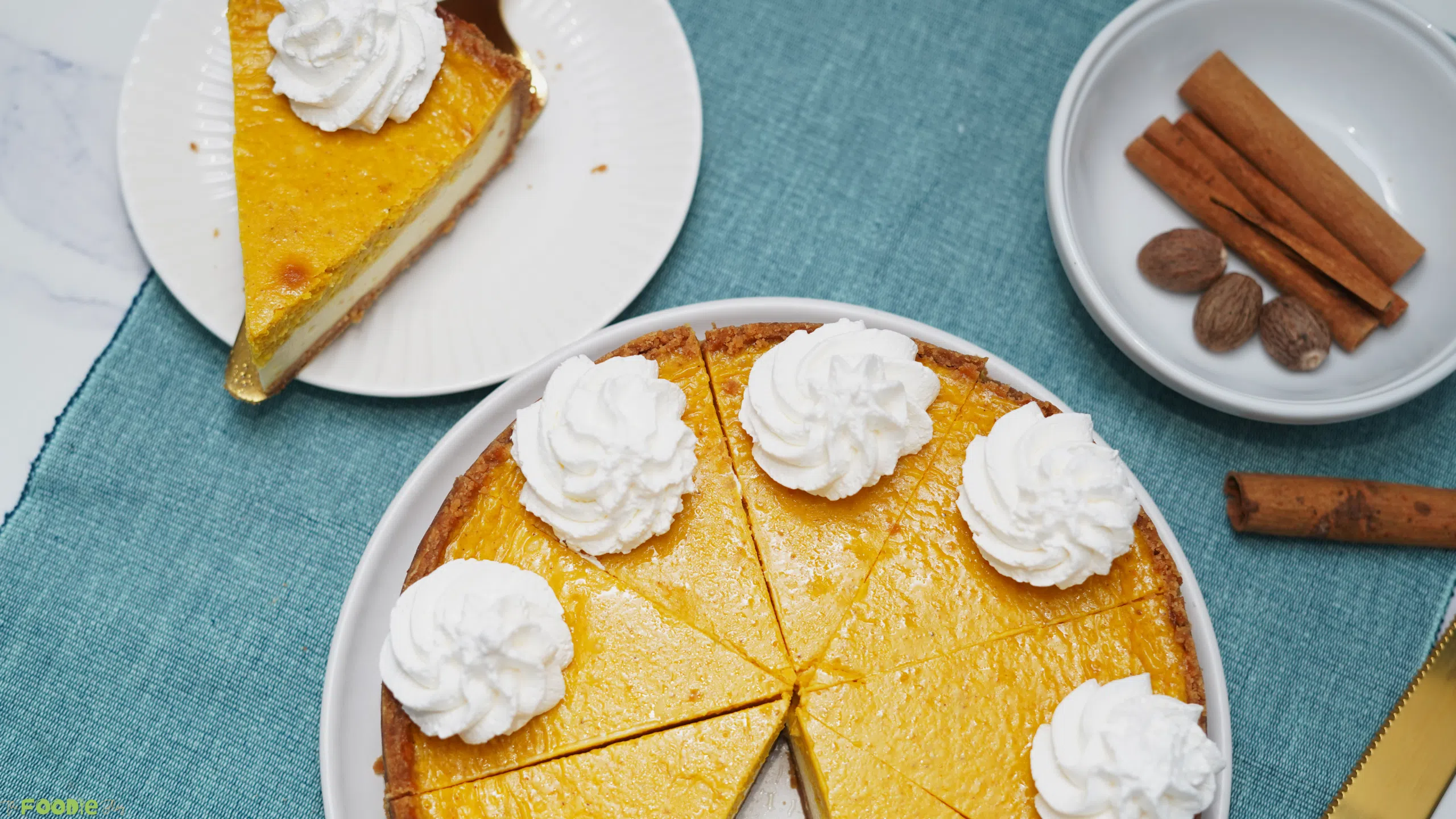 A sliced pumpkin pie cheesecake topped with whipped cream on a white plate with cinnamon sticks and nutmeg nearby.
