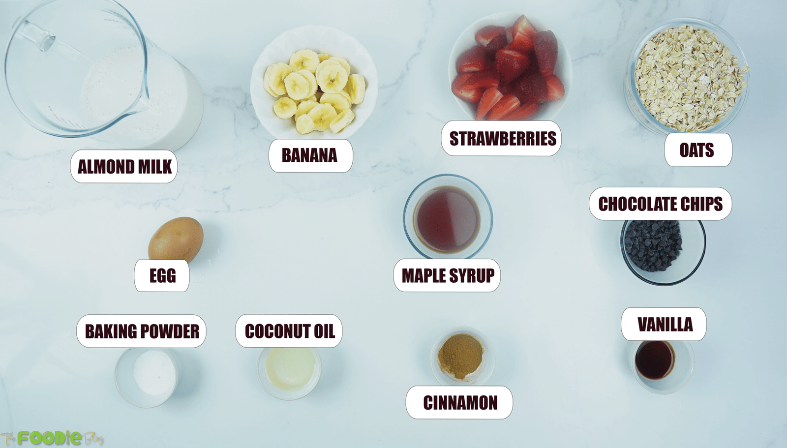 Measured ingredients for strawberry banana baked oatmeal including almond milk, banana, strawberries, oats, egg, maple syrup, chocolate chips, vanilla, baking powder, coconut oil, and cinnamon