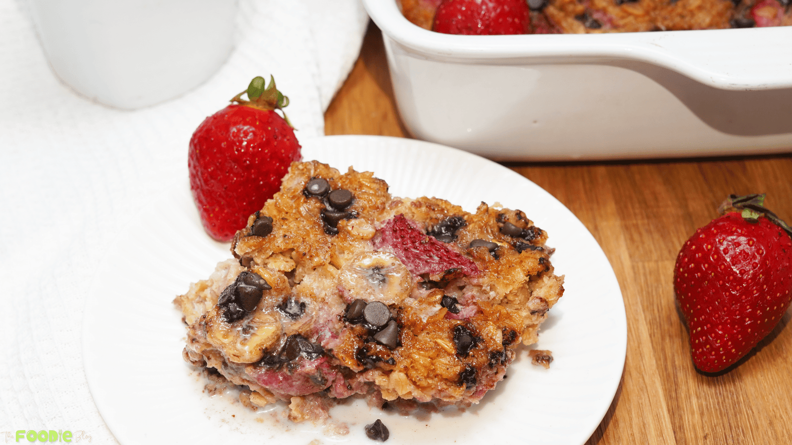Plated serving of strawberry banana baked oatmeal with fresh strawberries on a wooden board and the baking dish in the background