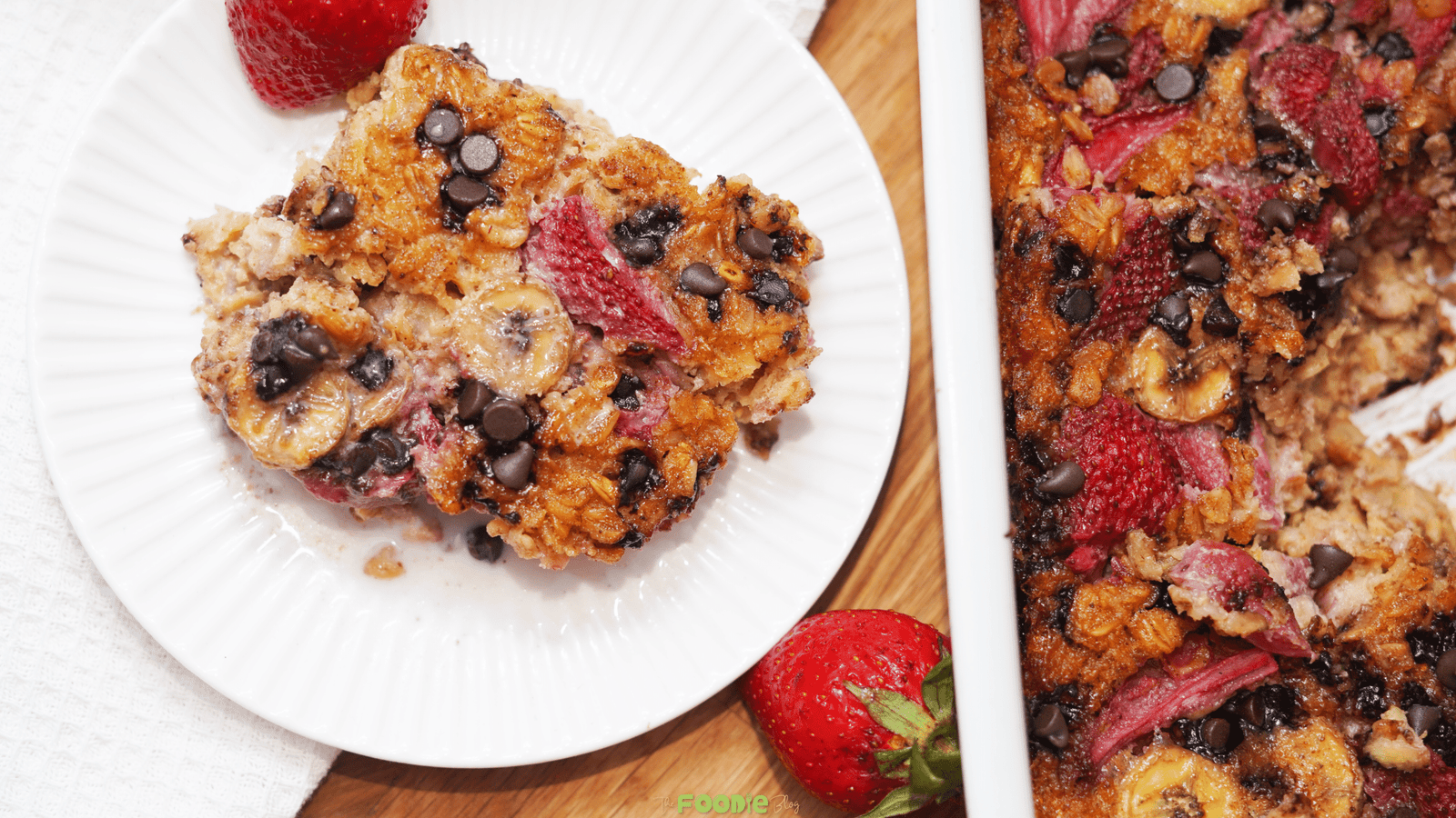 Overhead view of a serving of strawberry banana baked oatmeal on a white plate next to the baking dish and fresh strawberries