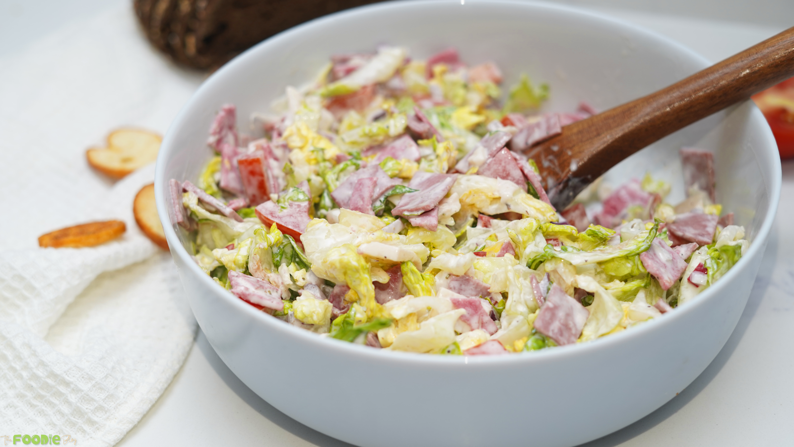 Hoagie dip mixed in a white bowl with chopped salami, pepperoni, lettuce, tomato, red onion, provolone, and a wooden spoon.