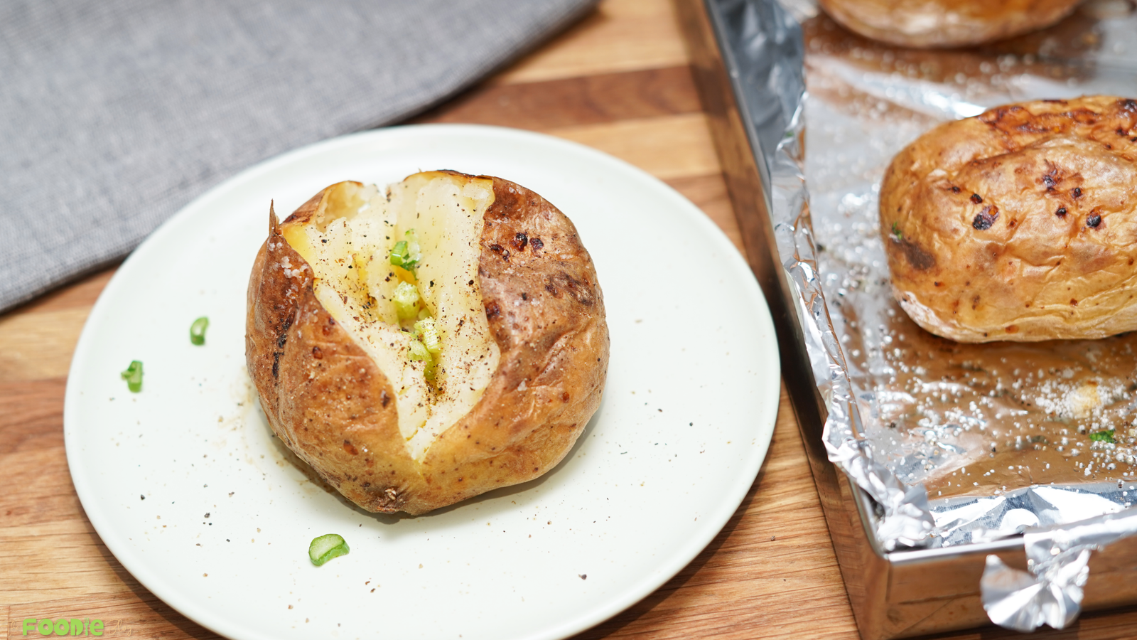 A baked russet potato opened on a white plate with seasoning and green onion, with more baked potatoes on a tray nearby.