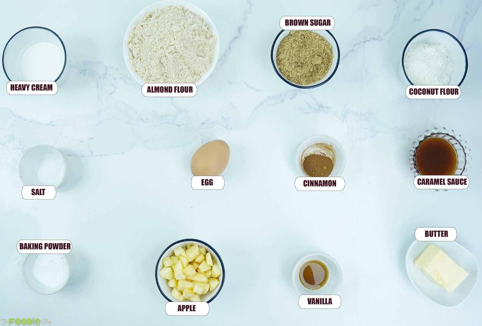 Ingredients for keto caramel apple scones arranged on a marble surface, including almond flour, coconut flour, apple, butter, cinnamon, and caramel sauce.