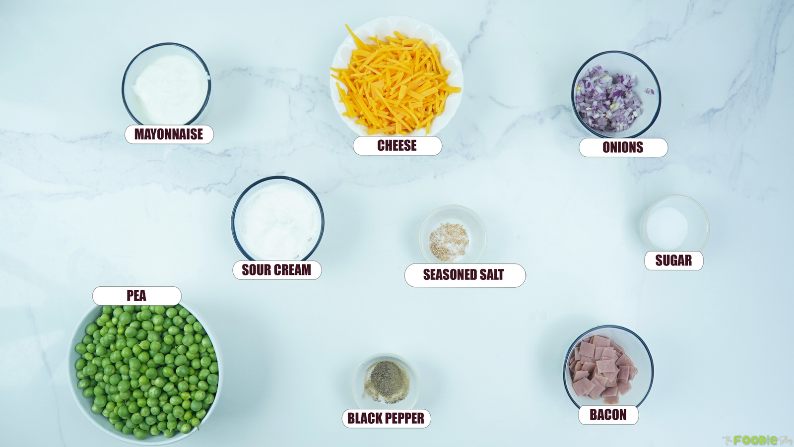 Ingredients for pea salad arranged on a marble surface, including peas, mayonnaise, sour cream, cheese, onions, bacon, sugar, seasoned salt, and black pepper.