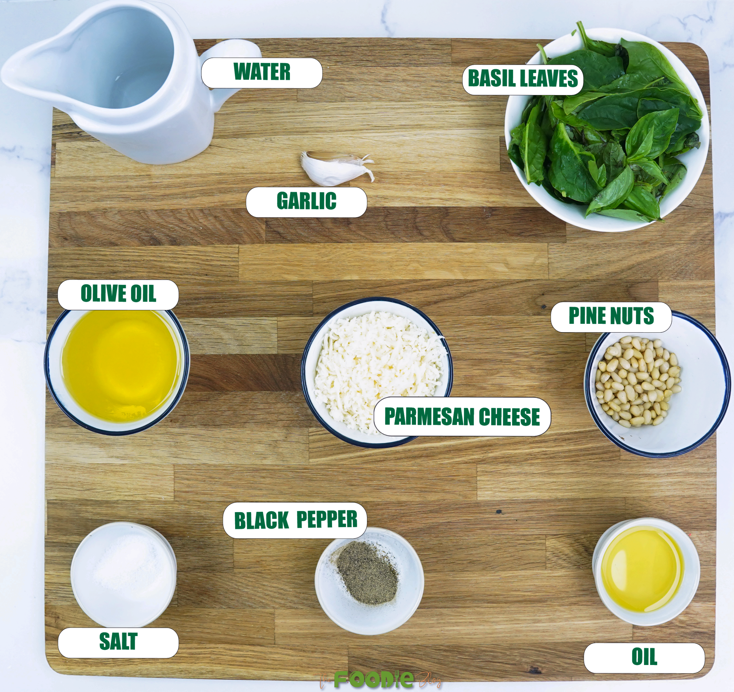 ingredients for pesto pasta arranged on a wooden board including basil, pine nuts, Parmesan, garlic, olive oil, salt, pepper, and water