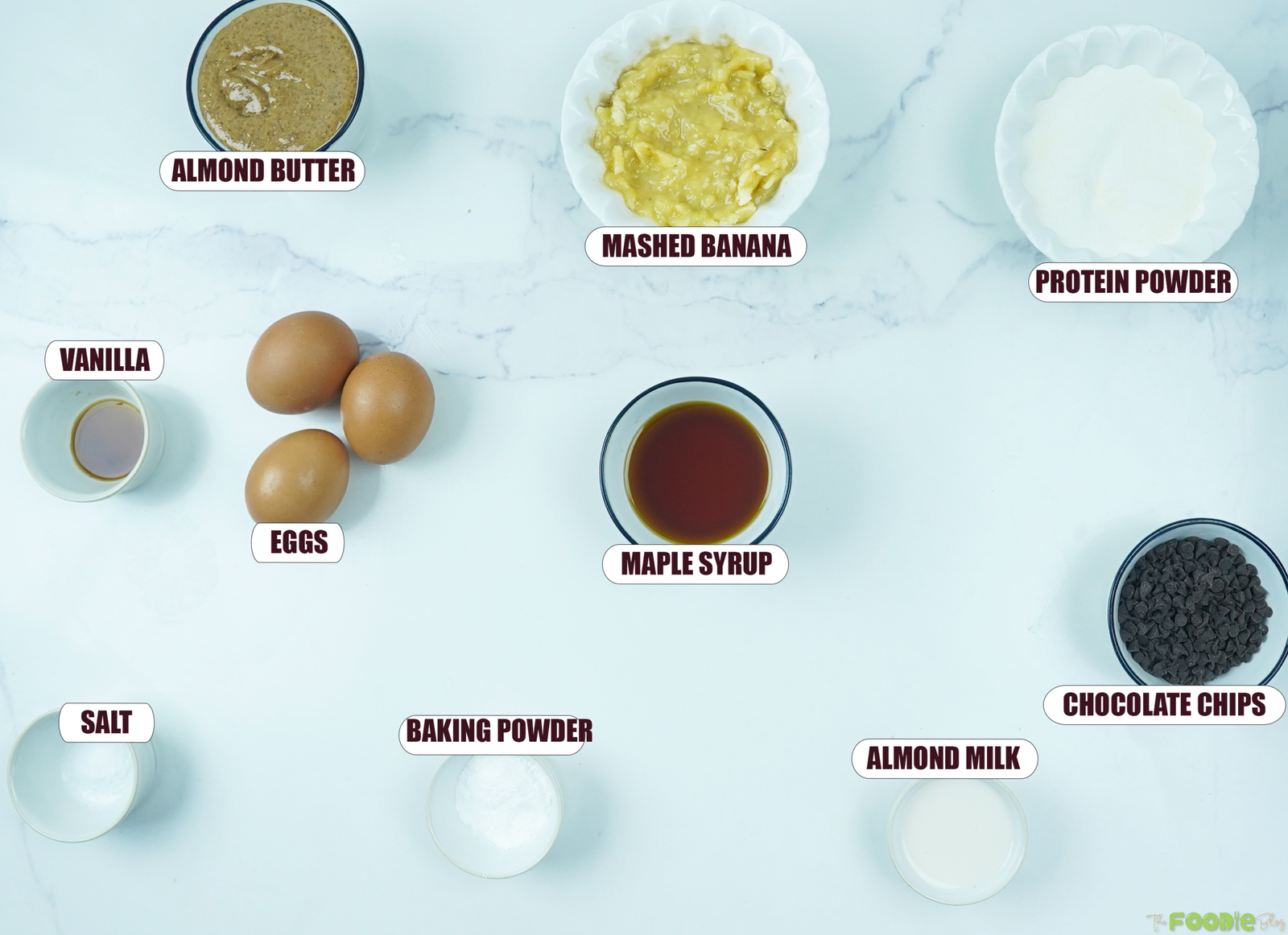 Overhead view of ingredients for protein muffins including banana, almond butter, eggs, protein powder, maple syrup, almond milk, and chocolate chips.
