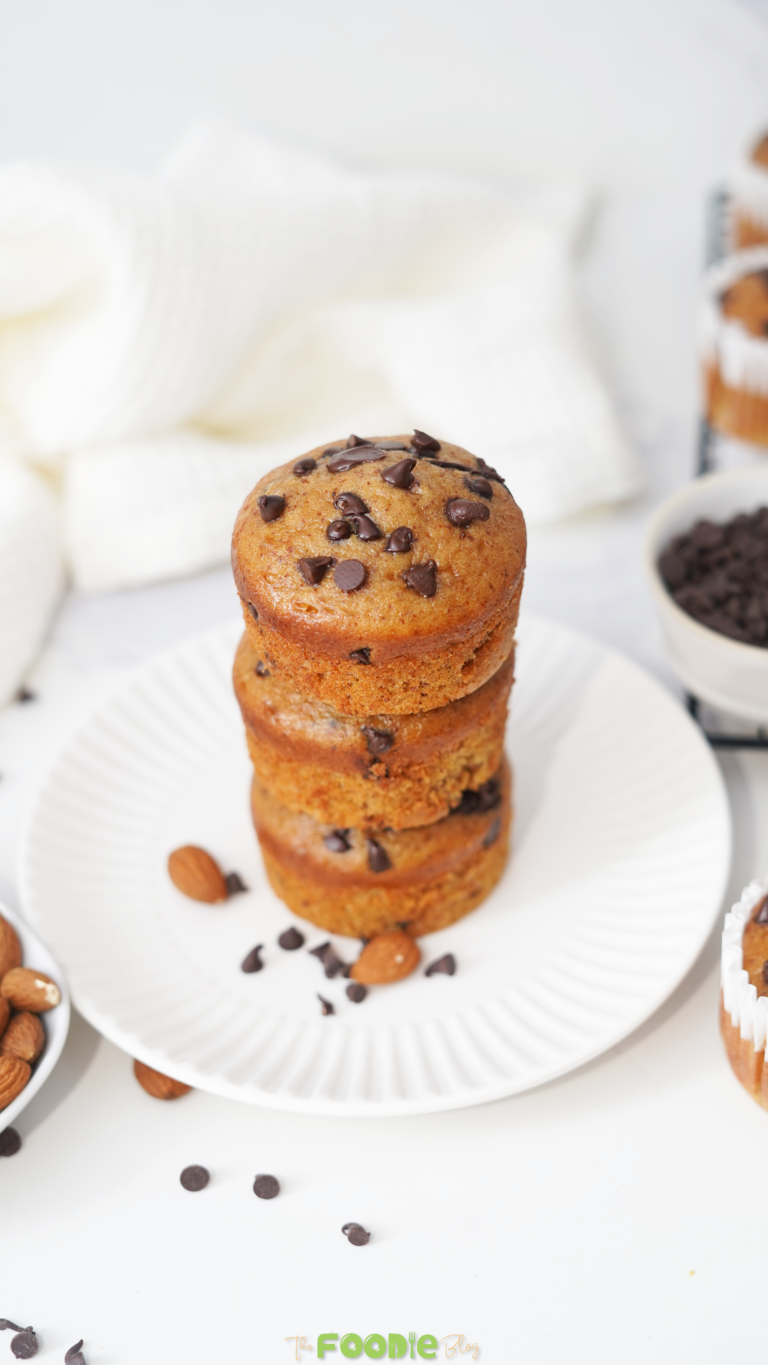 Three chocolate chip protein muffins stacked on a white plate with almonds and chocolate chips around them.