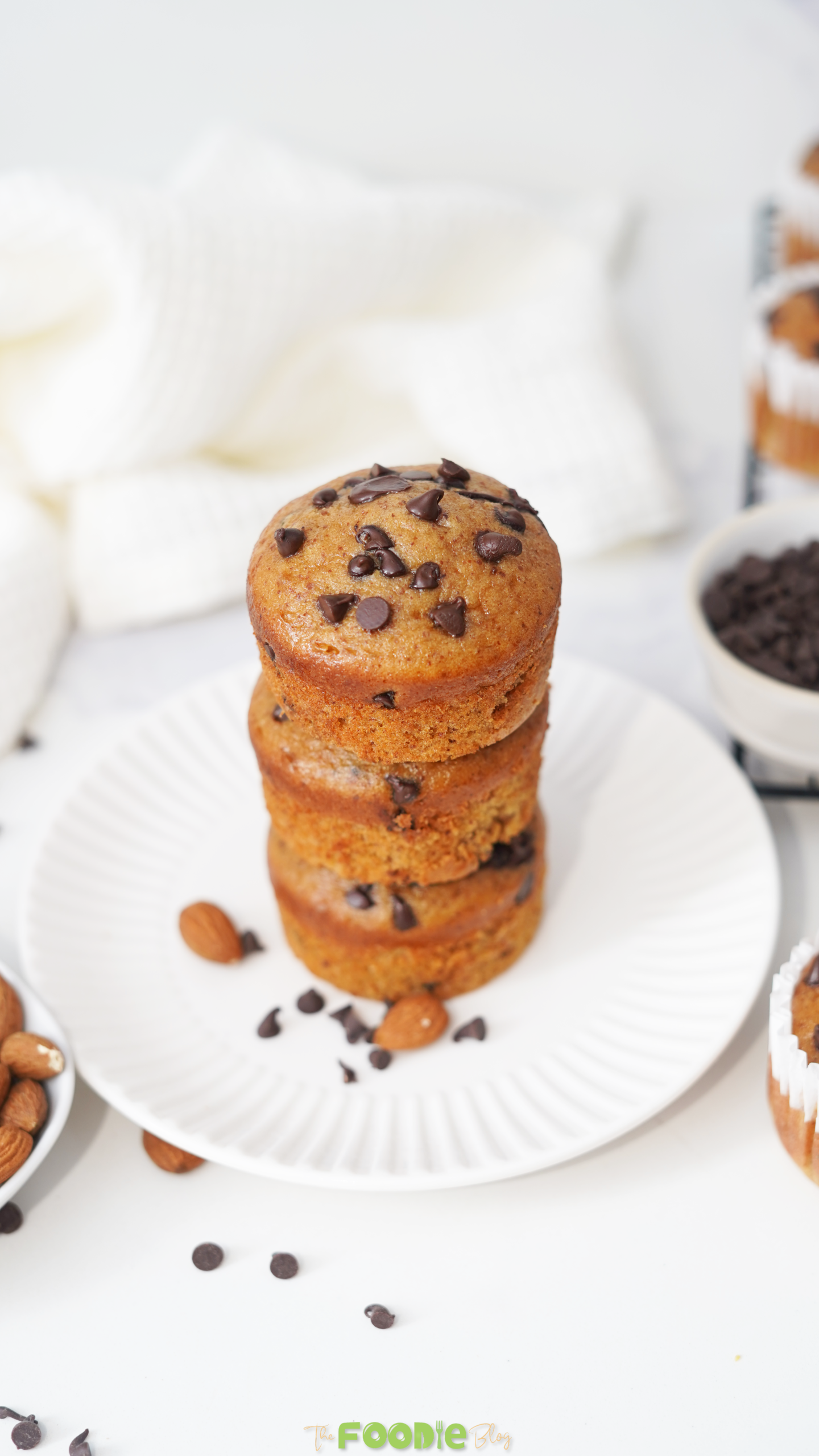 Three chocolate chip protein muffins stacked on a white plate with almonds and chocolate chips around them.
