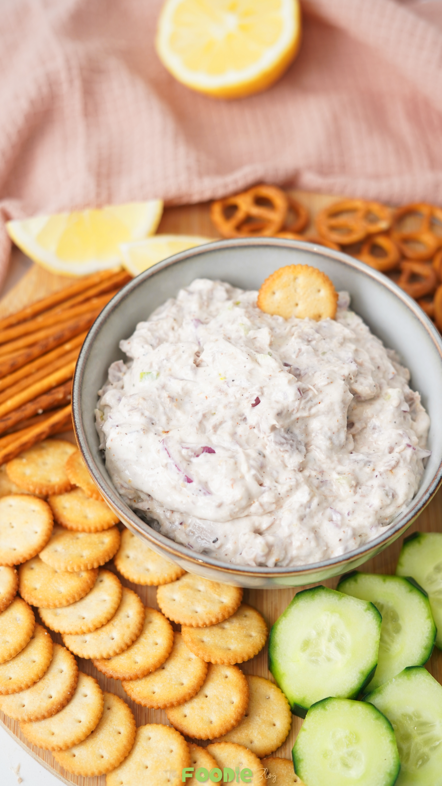 Close-up of creamy tuna dip in a bowl with round crackers, pretzel sticks, pretzels, sliced cucumbers, and lemon.