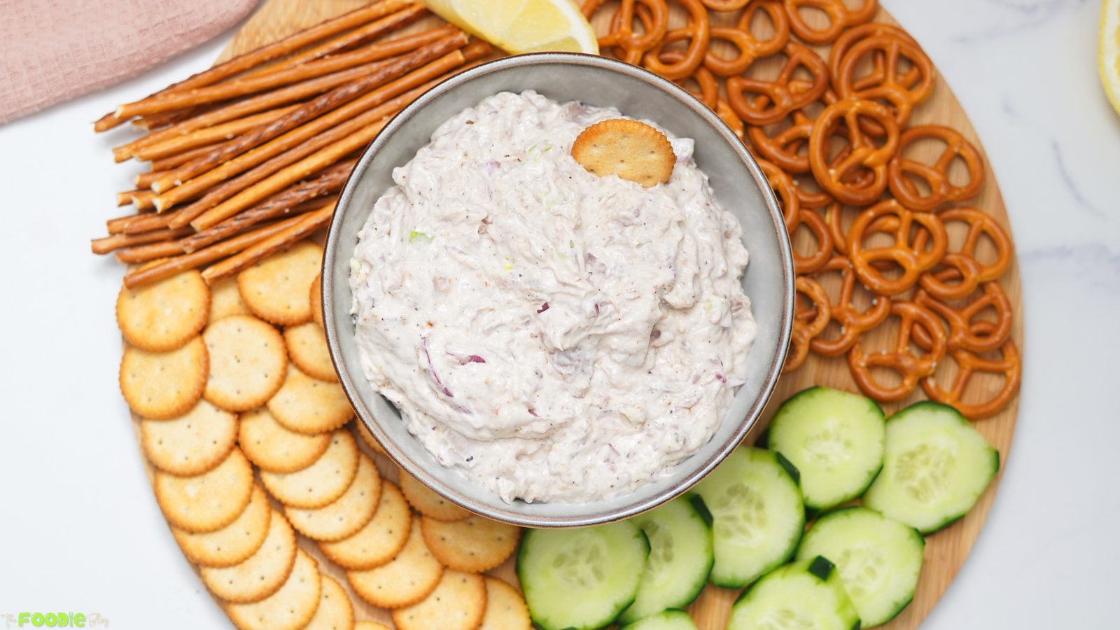 Overhead view of creamy tuna dip in a bowl with crackers, pretzel sticks, pretzels, cucumber slices, and lemon wedges on a wooden board.