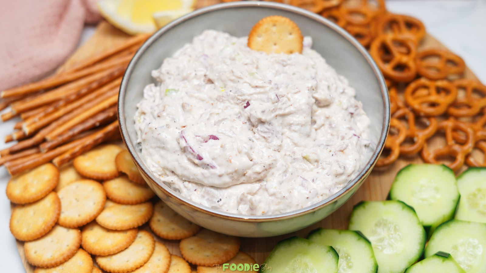 Bowl of creamy tuna dip served on a board with crackers, pretzel sticks, pretzels, cucumber slices, and lemon wedges.