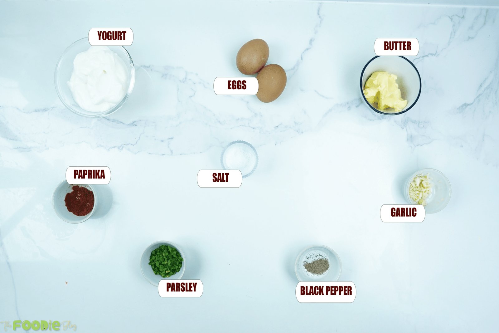Overhead view of ingredients for Turkish eggs including yogurt, eggs, butter, garlic, herbs, paprika, salt, and black pepper