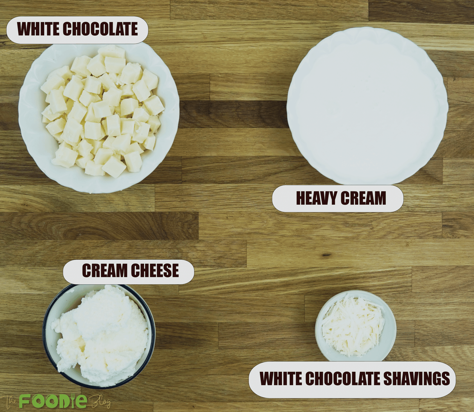 Ingredients for white chocolate mousse including white chocolate, heavy cream, cream cheese, and white chocolate shavings on a wooden surface