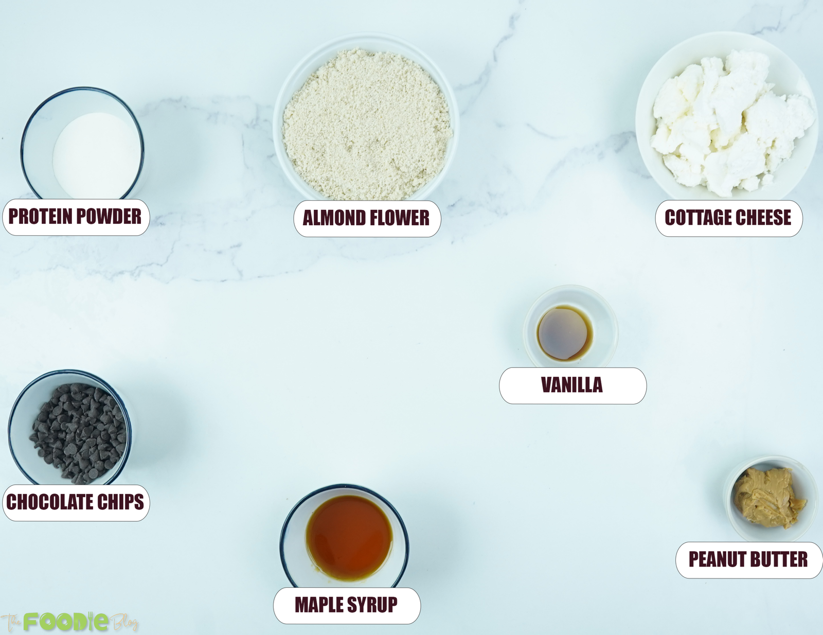 Ingredients for cottage cheese cookie dough arranged on a white surface, including cottage cheese, almond flour, protein powder, maple syrup, vanilla, peanut butter, and chocolate chips.