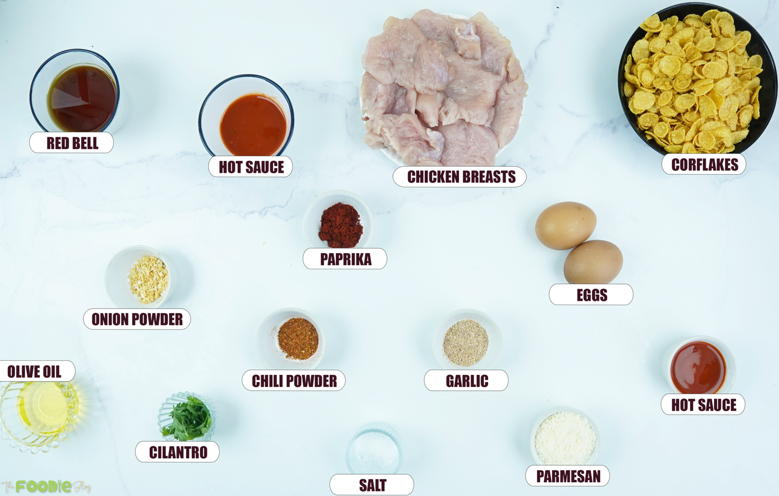 Ingredients for baked crunchy hot honey chicken arranged on a white surface with chicken, cornflakes, eggs, spices, Parmesan, honey, hot sauce, and herbs.