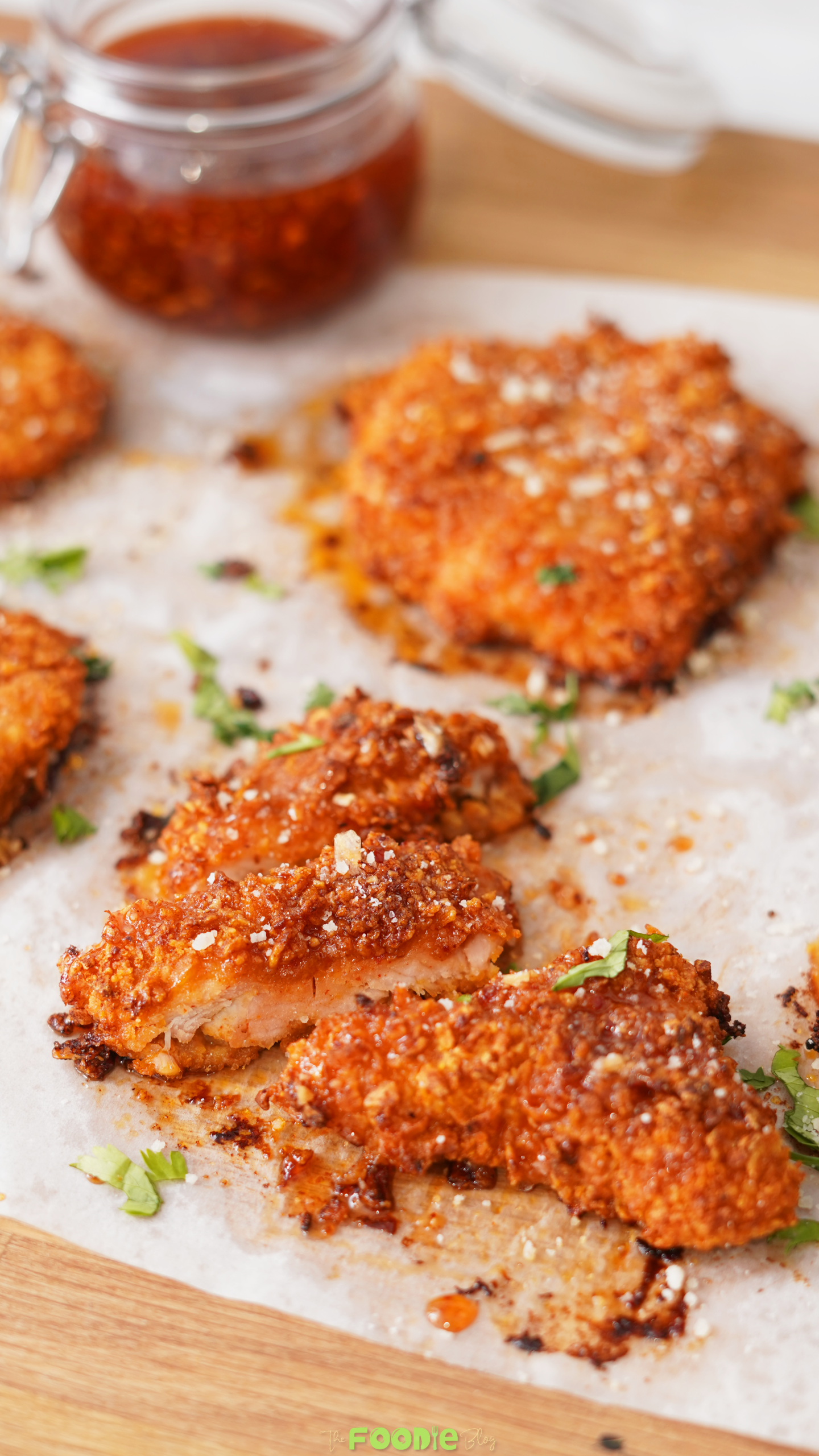 Crispy hot honey chicken pieces on parchment paper with fresh herbs, Parmesan, and a jar of spicy honey sauce in the background.