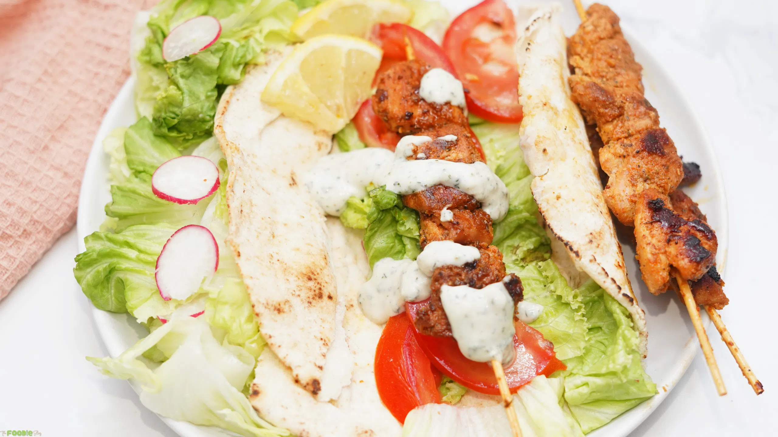 Chicken skewer served in pita bread with lettuce, tomato, radish, and creamy sauce on a white plate.