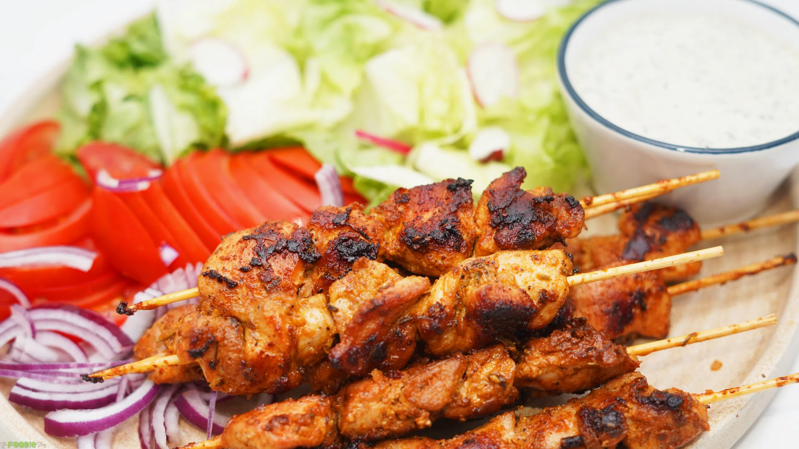 Charred chicken skewers served on a platter with lettuce, tomatoes, red onion, pita bread, lemon, and creamy sauce.
