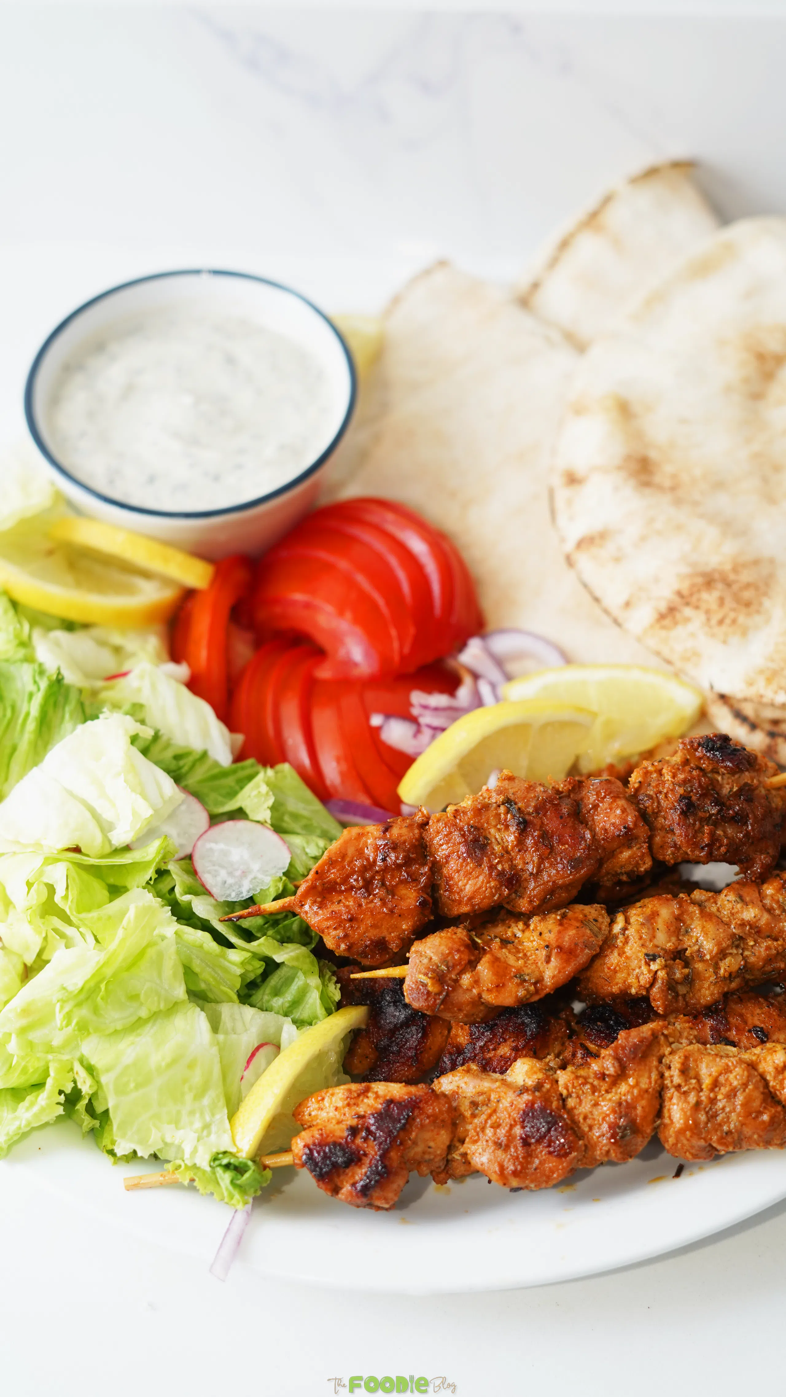 Chicken skewers served with lettuce, tomato, red onion, lemon wedges, pita bread, and creamy yogurt sauce.