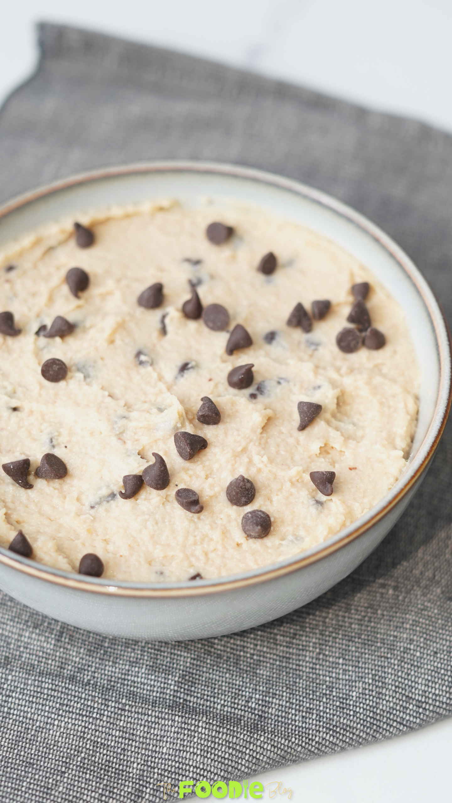Close-up of creamy cottage cheese cookie dough topped with chocolate chips in a shallow bowl on a gray cloth.