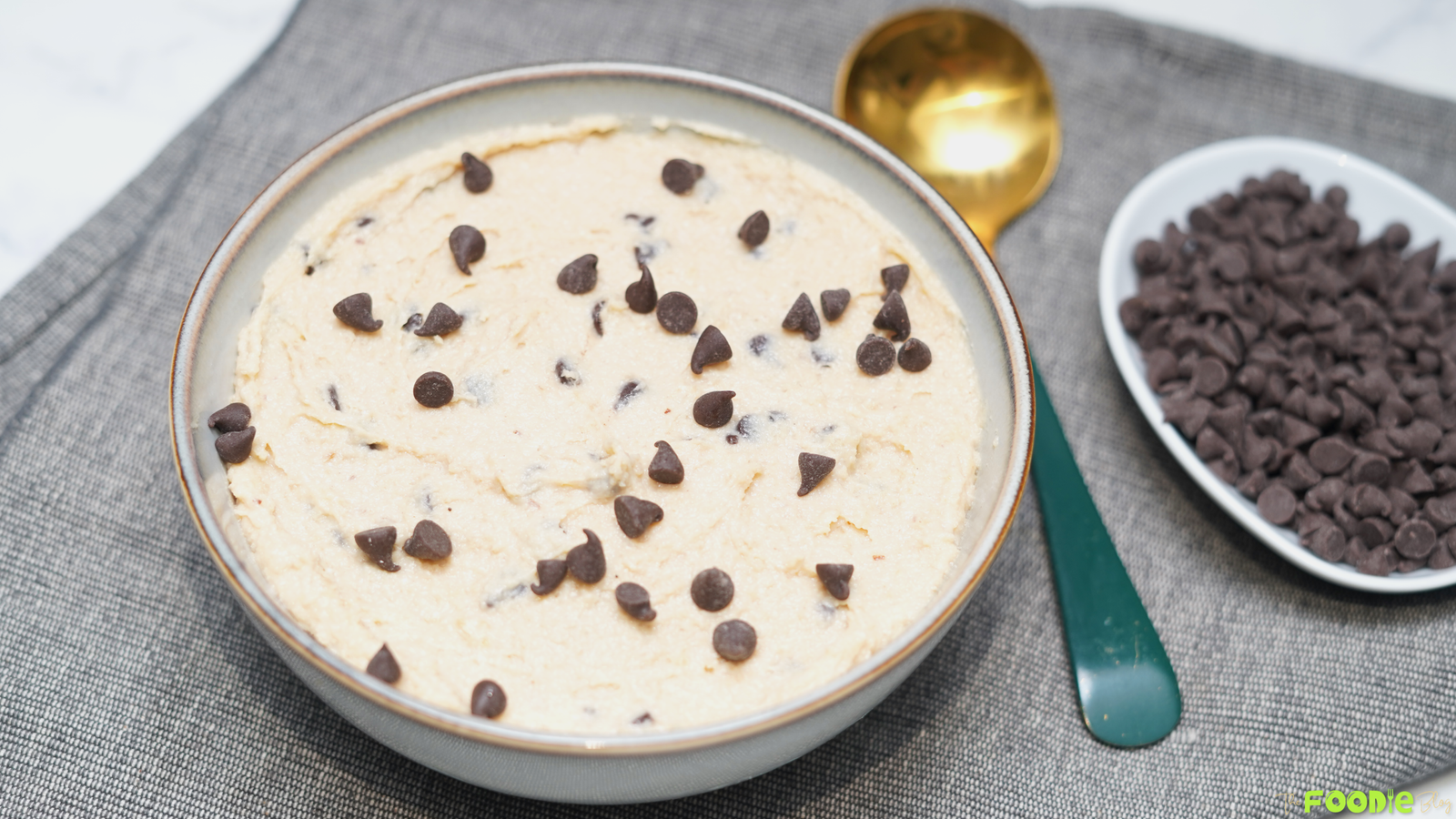 Creamy cottage cheese cookie dough in a bowl topped with chocolate chips, served with a spoon and extra chocolate chips on the side.