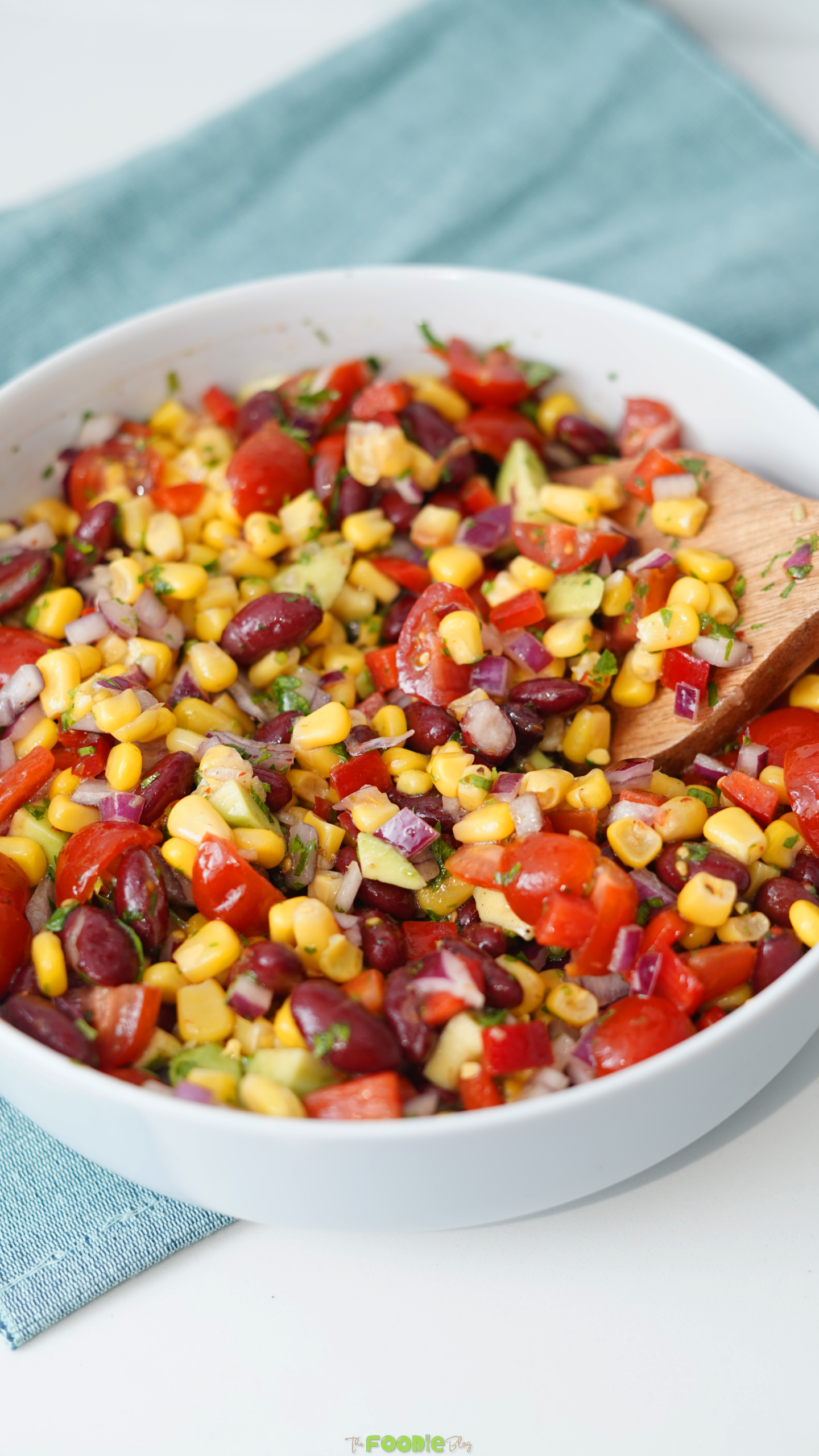 Cowboy bean salad in a white bowl with corn, beans, tomatoes, red onion, avocado, cilantro, red bell pepper, and a wooden spoon.