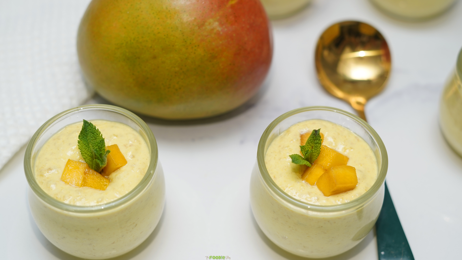 Two glass cups of mango mousse topped with mango cubes and mint on a bright marble surface.