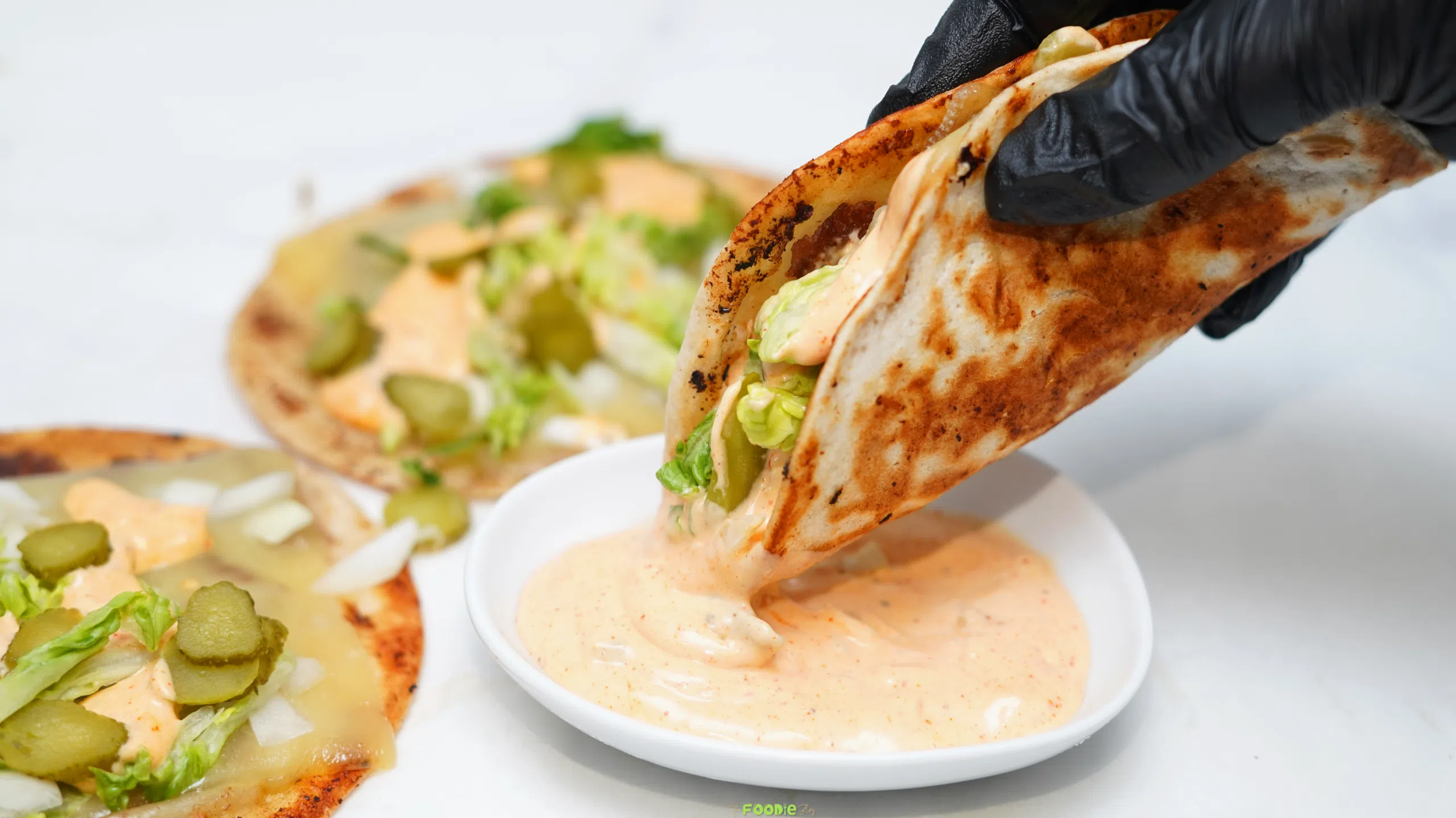 A folded smash burger taco being dipped into creamy burger sauce with more tacos in the background.