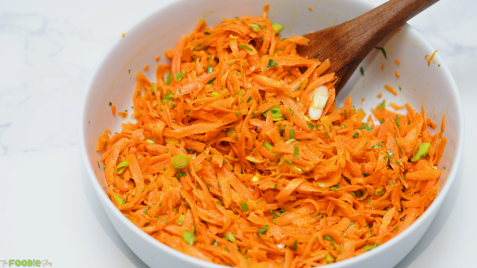 Shredded carrot salad tossed with green onion, parsley, and honey Dijon dressing in a white bowl with a wooden spoon.