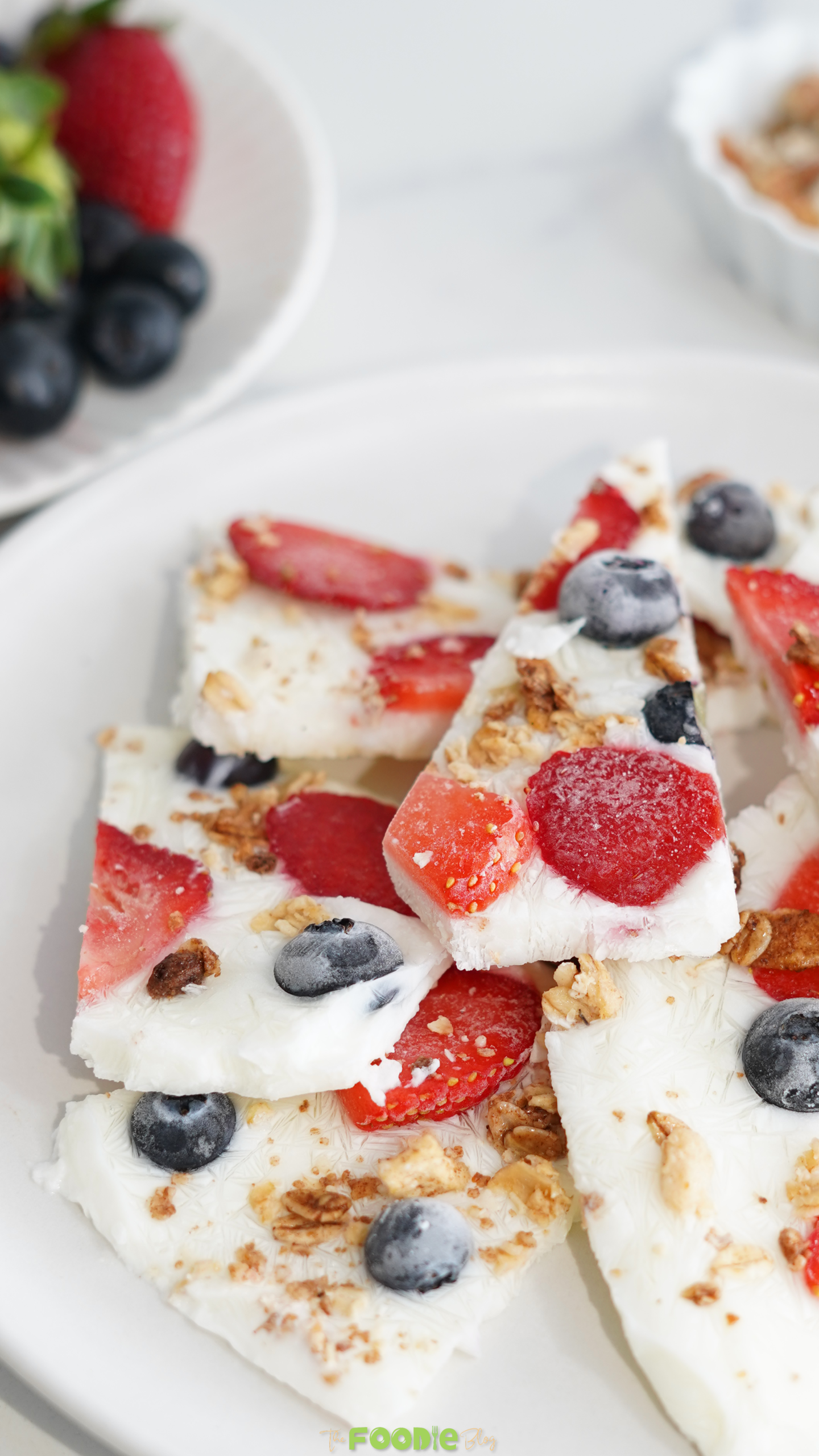 Pieces of frozen yogurt bark topped with strawberries, blueberries, and granola on a white plate.