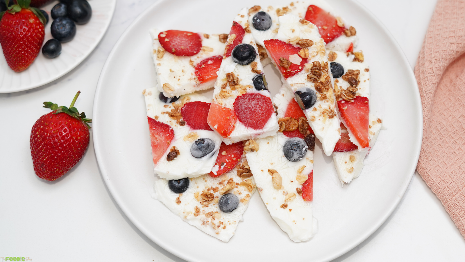 Frozen yogurt bark pieces served on a white plate with strawberries, blueberries, and a pink towel nearby.