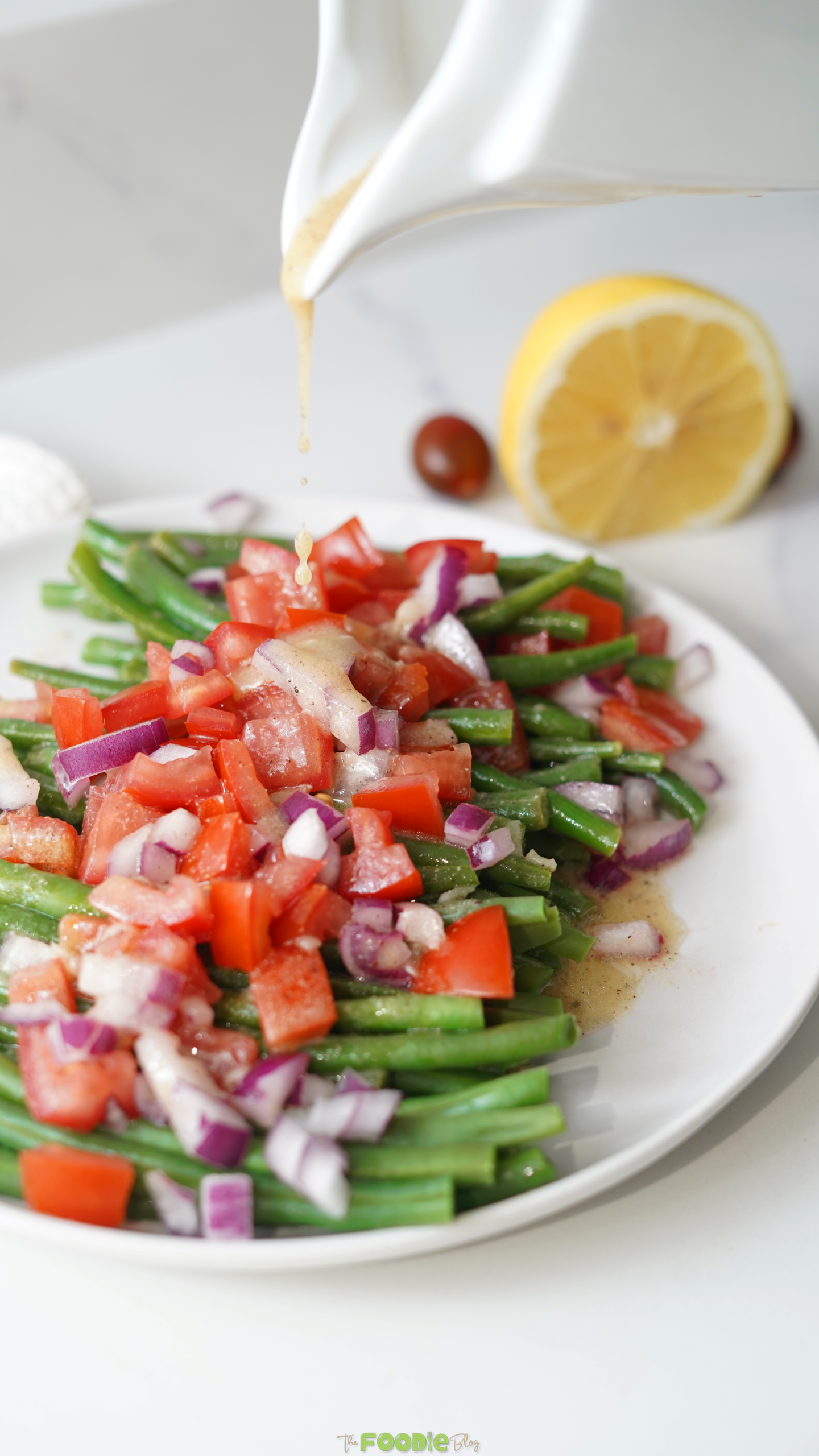 Fresh green bean salad with diced tomato and red onion served on a white plate with lemon on the side.