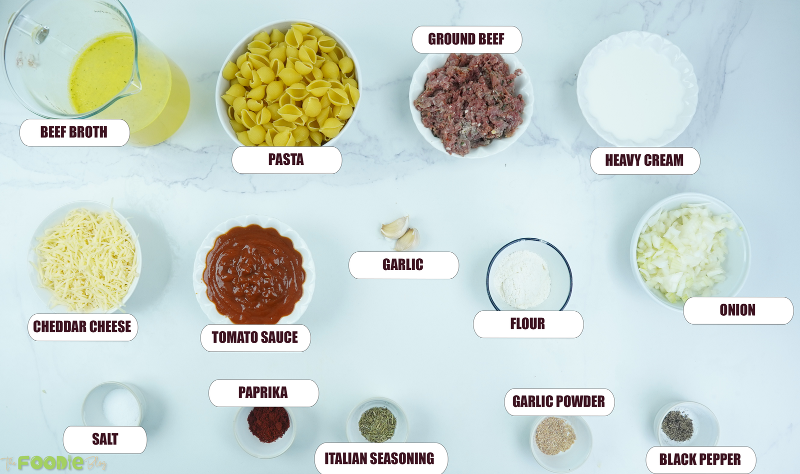 Ingredients for Homemade Hamburger Helper arranged on a white marble surface, including pasta shells, ground beef, beef broth, tomato sauce, cheddar cheese, onion, garlic, cream, flour, and seasonings.