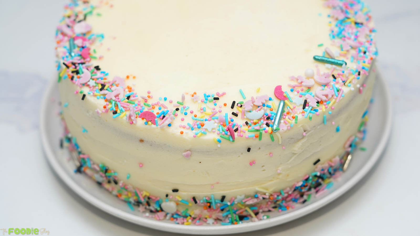 Close-up of a frosted vanilla cake decorated with colorful rainbow sprinkles around the top edge.