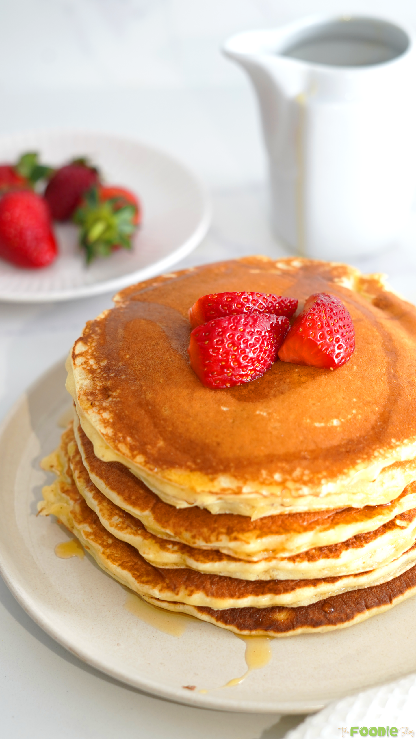Tall stack of golden protein pancakes with strawberries, syrup, and a white pitcher in the background.