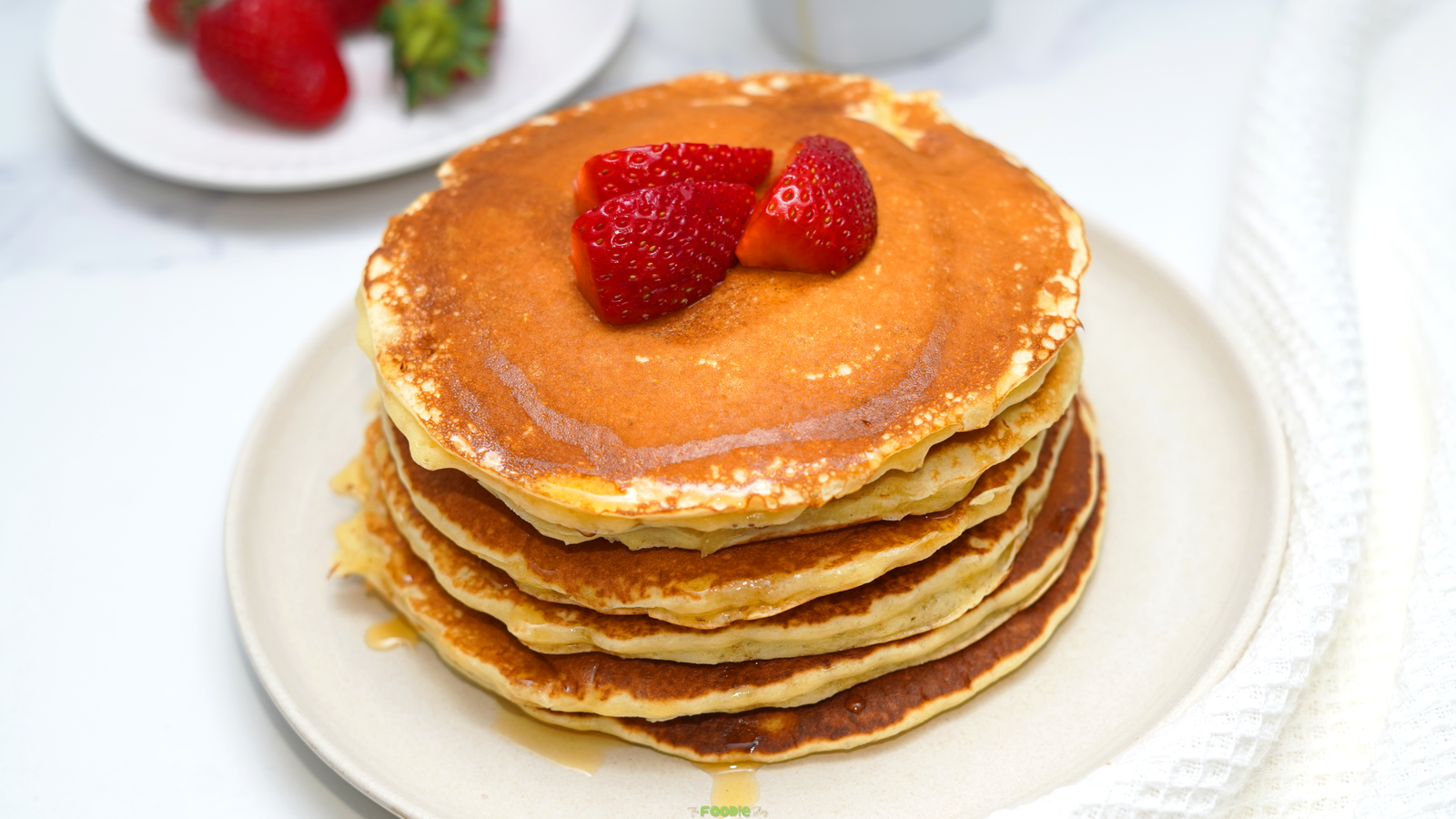Stack of golden protein pancakes topped with fresh strawberries and a drizzle of syrup on a white plate.