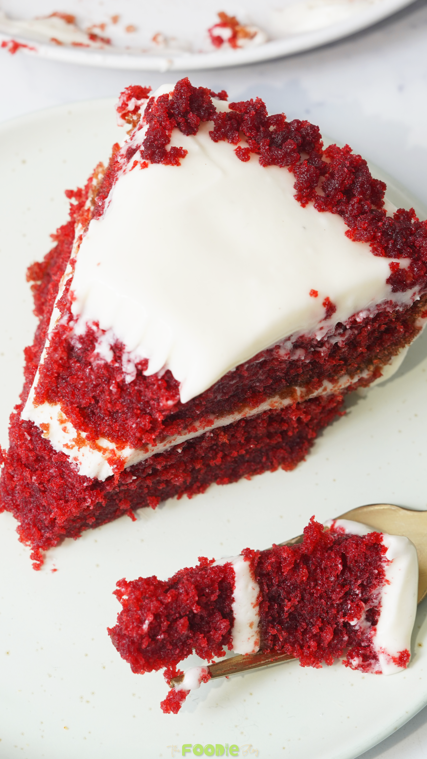 Close-up of a red velvet cake slice with cream cheese frosting, red crumbs, and a bite on a gold fork.
