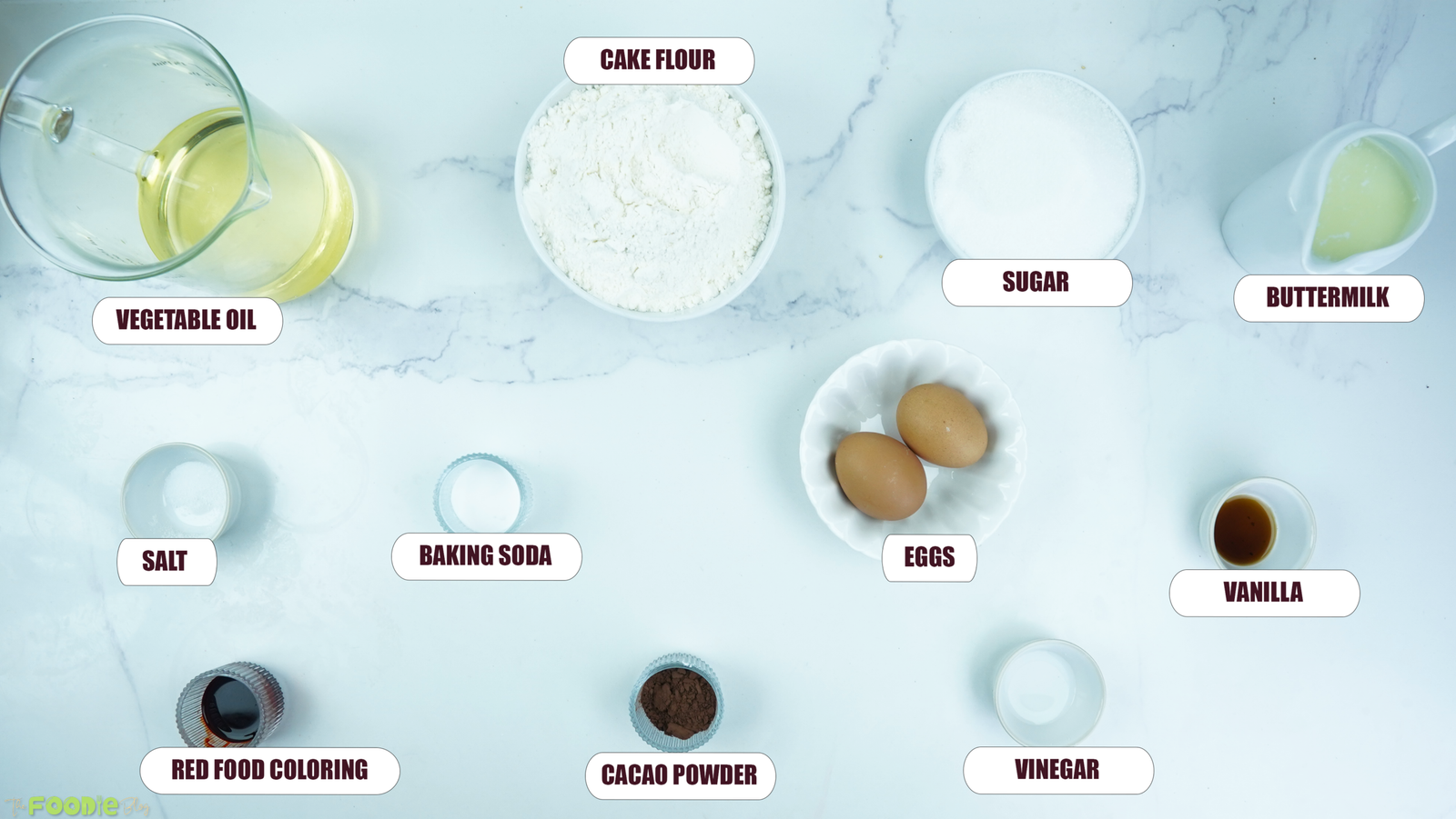 Ingredients for red velvet cake arranged on a white marble surface, including cake flour, sugar, buttermilk, eggs, cocoa powder, red food coloring, oil, vanilla, vinegar, baking soda, and salt.