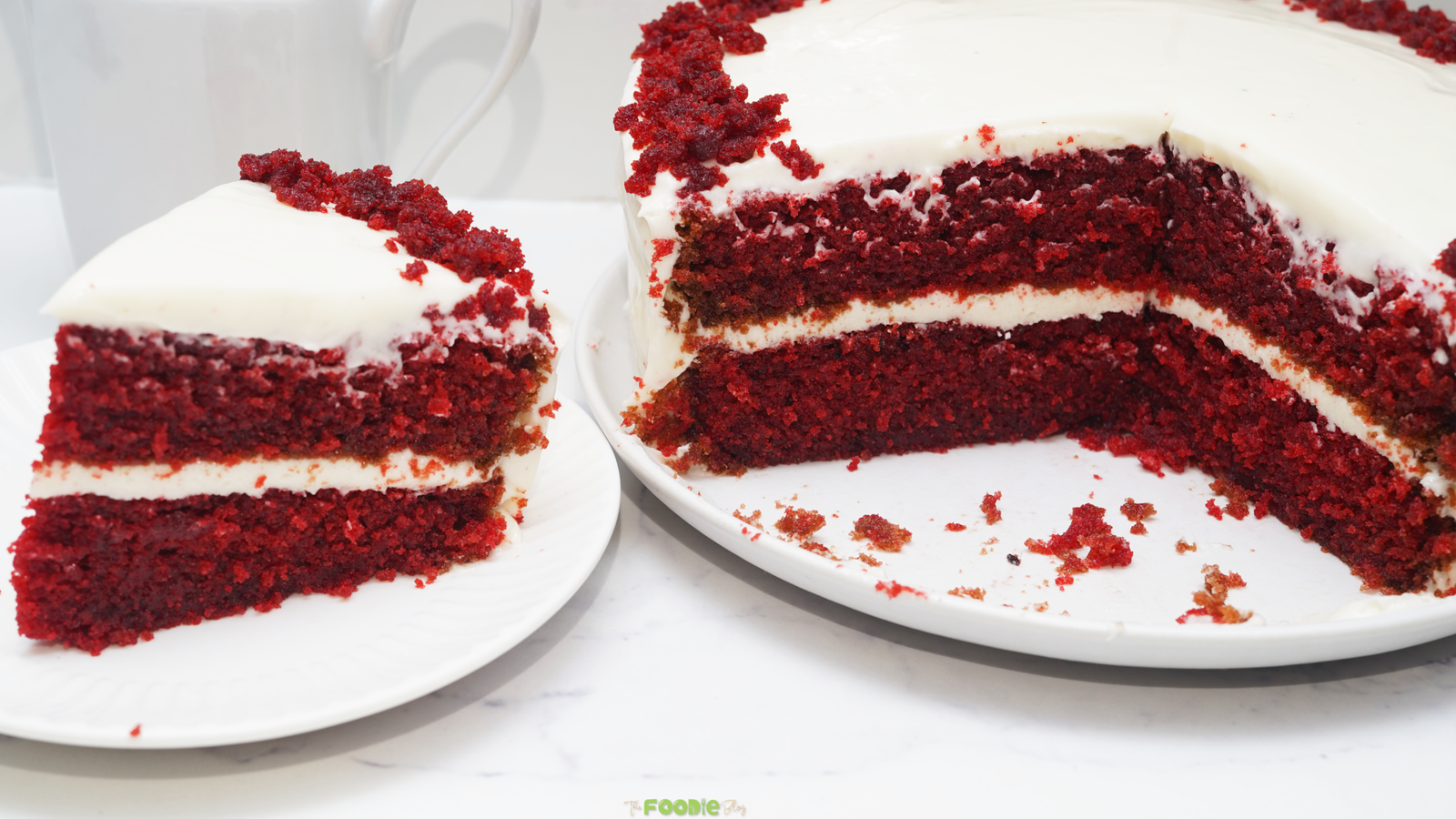 A sliced red velvet layer cake with cream cheese frosting and red cake crumbs on top, served beside a slice on a white plate.