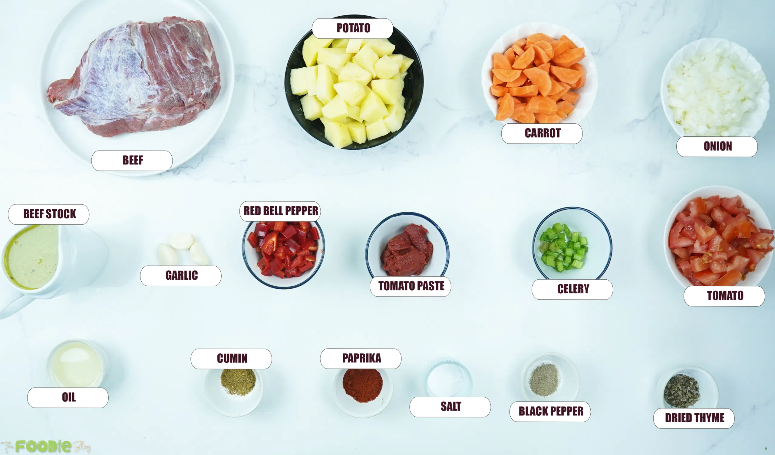 Ingredients for beef stew arranged on a white surface, including stewing beef, potatoes, carrots, onions, tomatoes, red bell pepper, celery, garlic, tomato paste, stock, and spices.