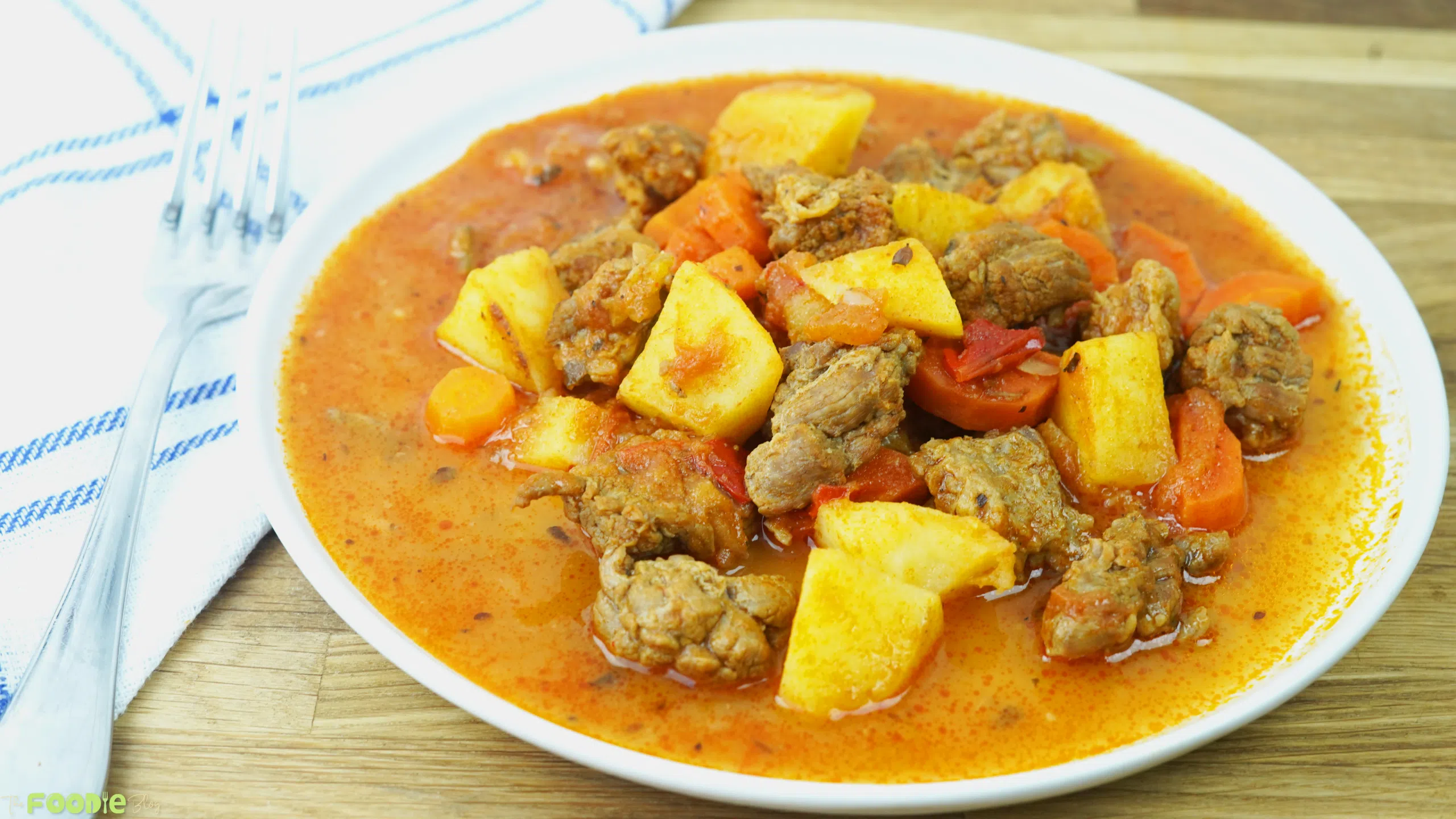 Beef stew served in a shallow white bowl with potatoes, carrots, beef chunks, and a red savory broth.