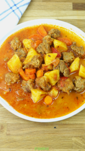 Bowl of beef stew with tender beef chunks, potatoes, carrots, and tomato broth served on a wooden board.