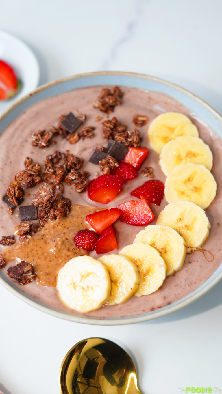 Close-up of a chocolate protein yogurt bowl with banana slices, strawberries, granola, chocolate chunks, and peanut butter.