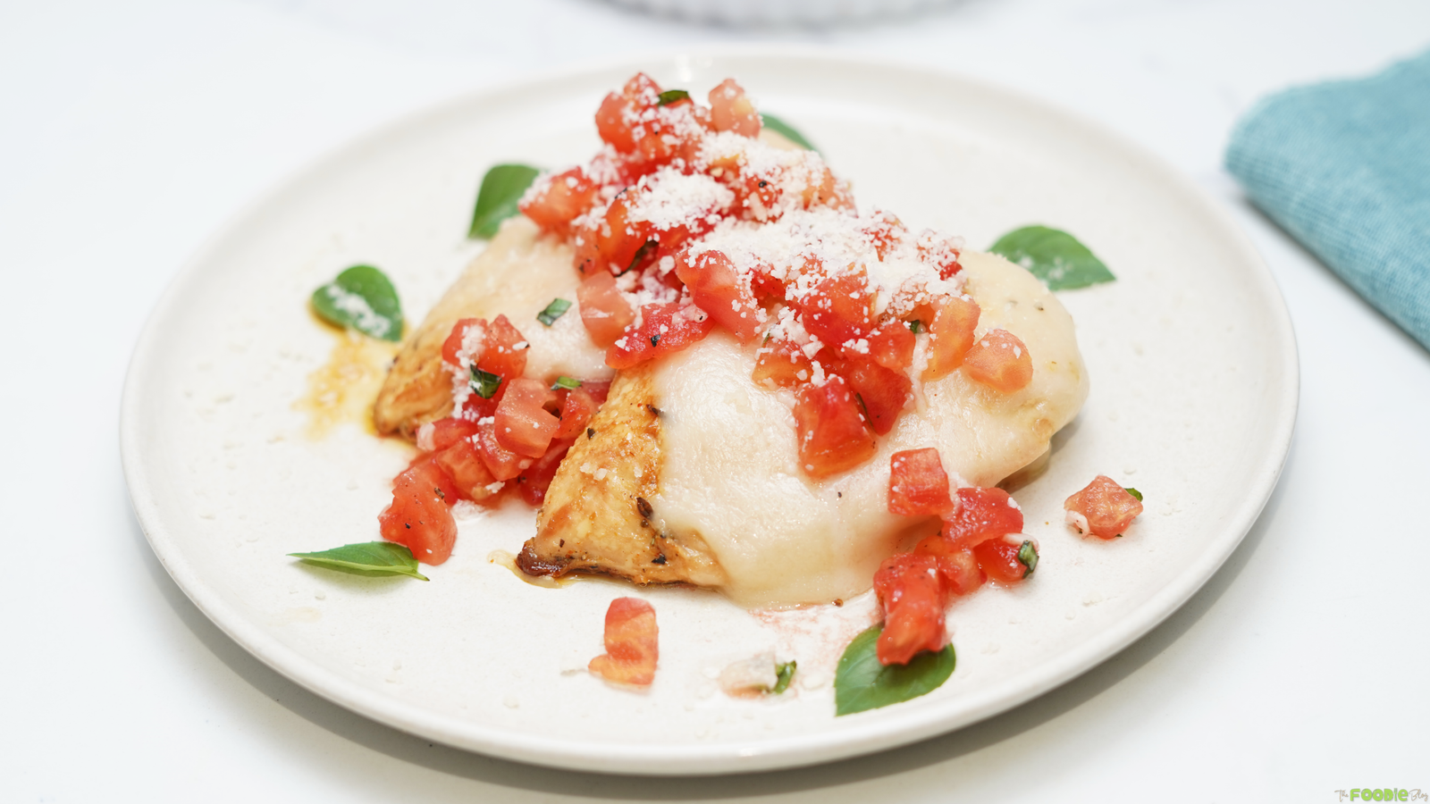 Bruschetta Chicken topped with melted mozzarella, chopped tomatoes, basil, and Parmesan on a white plate.