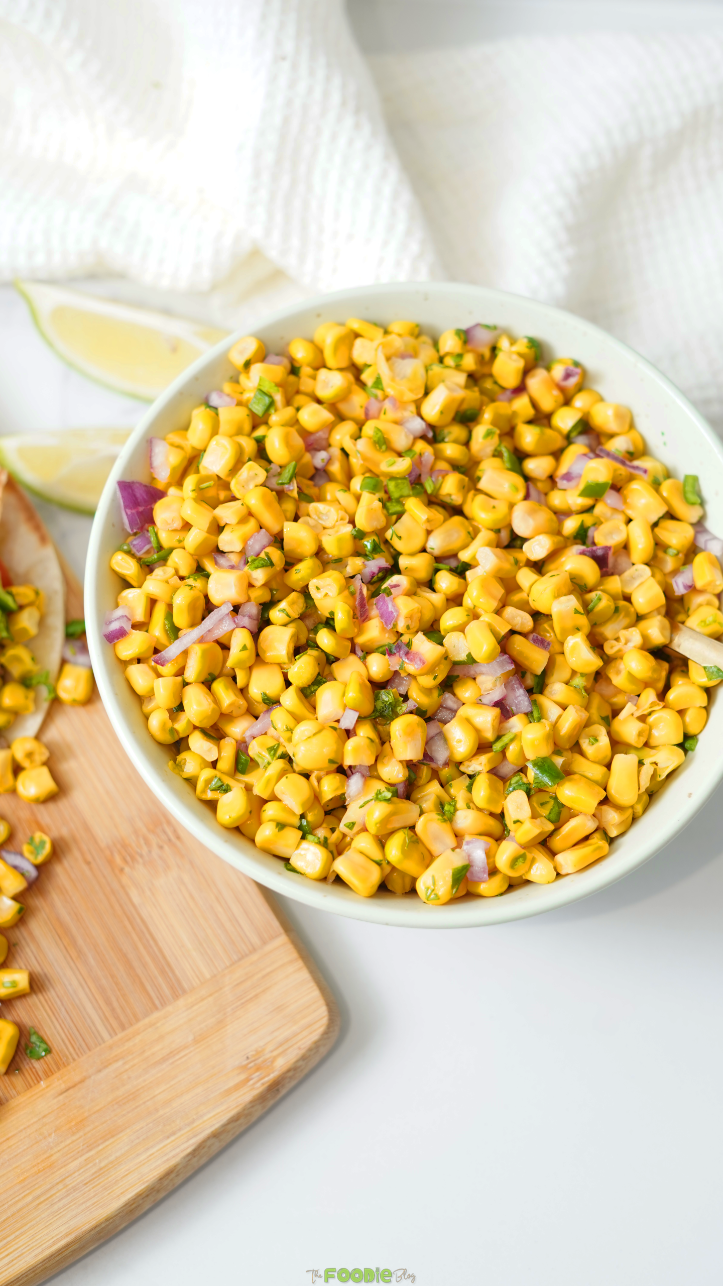 Bowl of fresh corn salsa with lime wedges and tacos in the background.