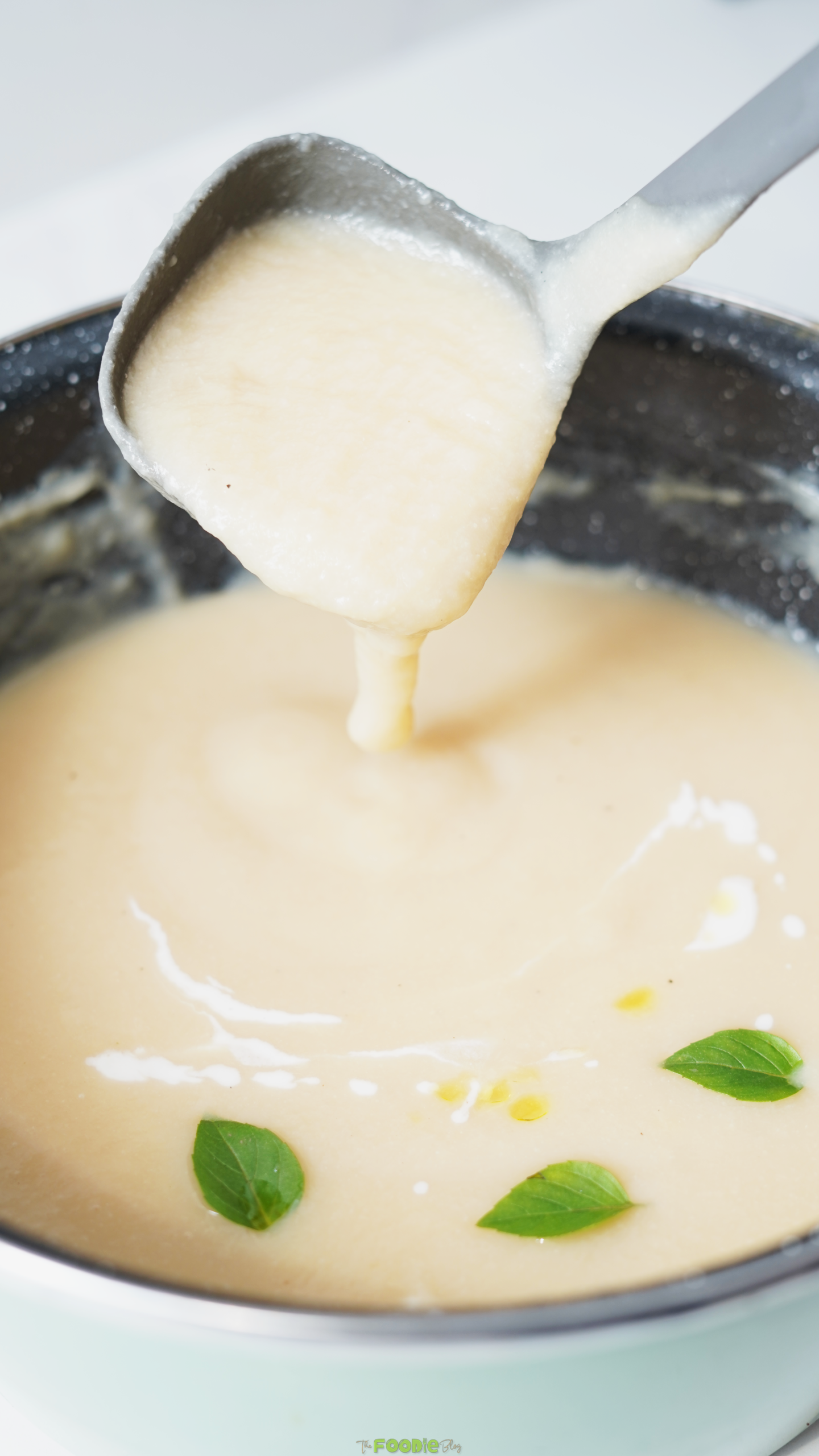 Ladle lifting smooth creamy cauliflower soup from a pot with fresh herb leaves on top.